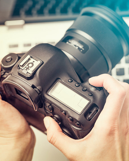 Two hands holding a modern digital DSLR camera with a computer in the background